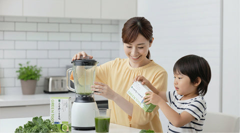 Mother and child in a bright kitchen preparing a green vegetable drink together, with the mother using a blender and the child holding a box of green juice powder on the counter surrounded by fresh greens.